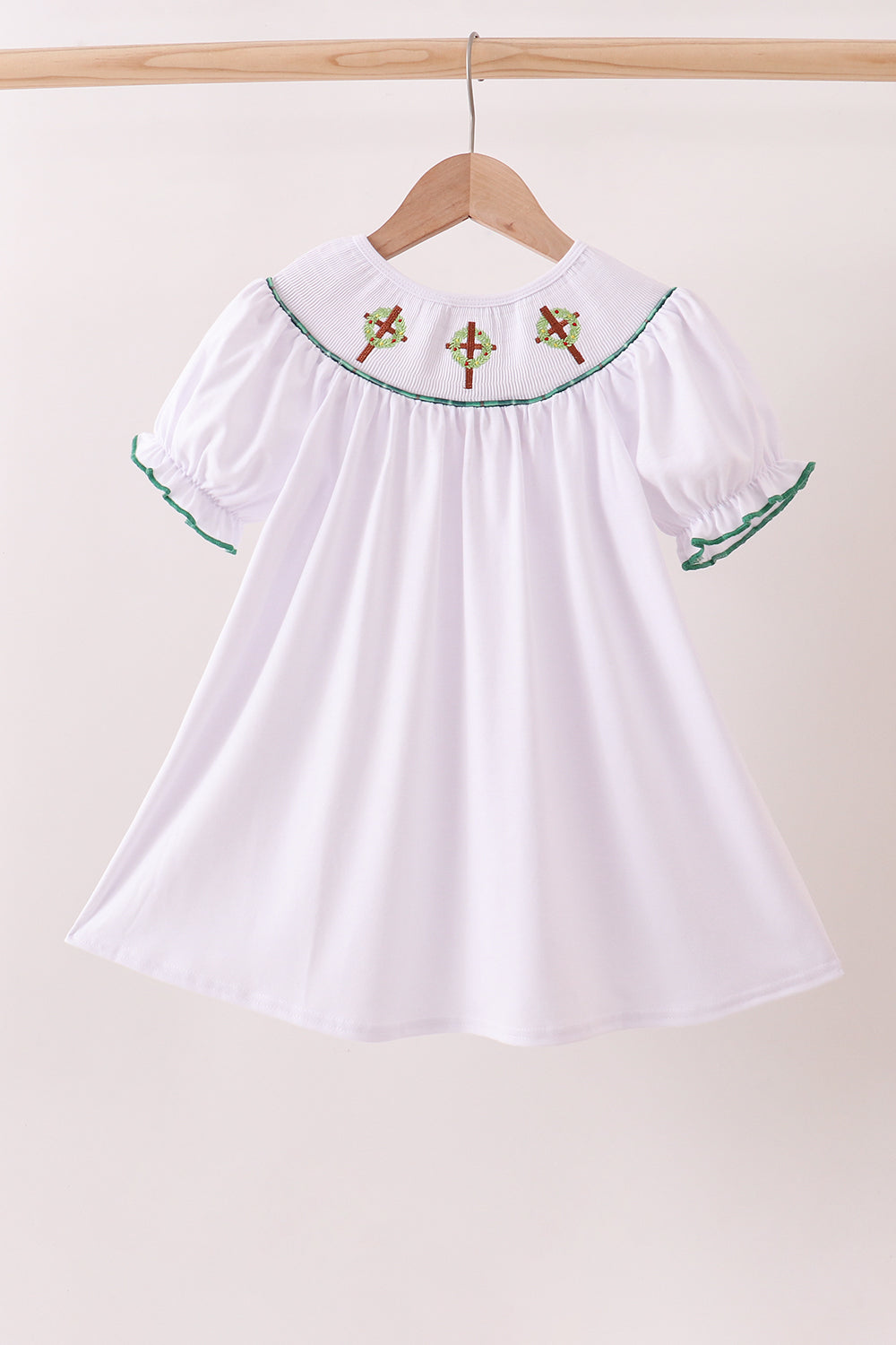 White dress with green trim and red embroidery on a hanger against a beige background Aaria Tees