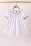 White dress with green trim and red embroidery on a hanger against a beige background Aaria Tees