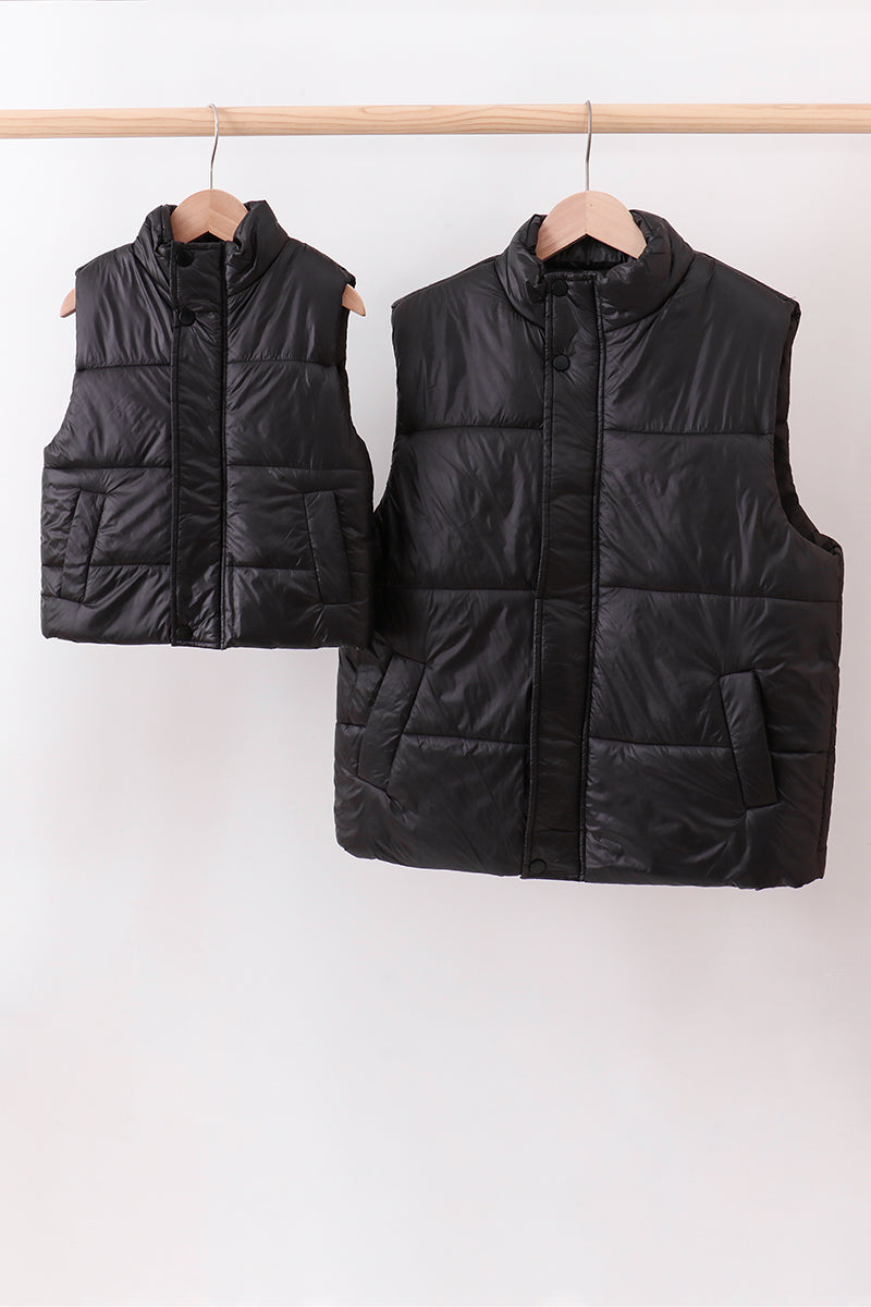 Two black puffer vests hanging on a rack against a white background Aaria Tees